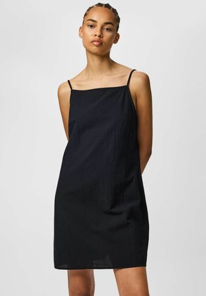 Kvinne med flettefrisyre iført en ermeløs svart kjole med tynne stropper, stående foran en ensfarget lys grå bakgrunn.