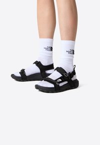 Zwarte sandalen voor buiten met verstelbare riemen, rubberen zolen en een gestructureerde afwerking, gedragen met witte sokken met een logo.