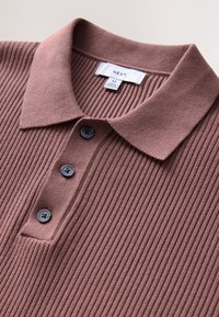 Geribd bruin poloshirt met een zachte textuur, een kraag en drie donkere knopen aan de voorkant. Het merklabelf geeft "NEXT" aan, maat M.