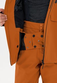 Orangene, wetterfeste Jacke mit schwarzem Futter, ausgestattet mit Druckknöpfen und einem Reißverschluss. Hose aus ähnlichem orangefarbenem Material und Akzenten.