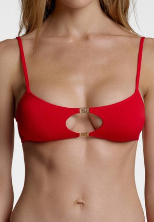 Kobieta nosząca czerwoną górę od bikini z cienkimi ramiączkami i centralnym wycięciem, trzymanym przez dwa złote klipsy.
