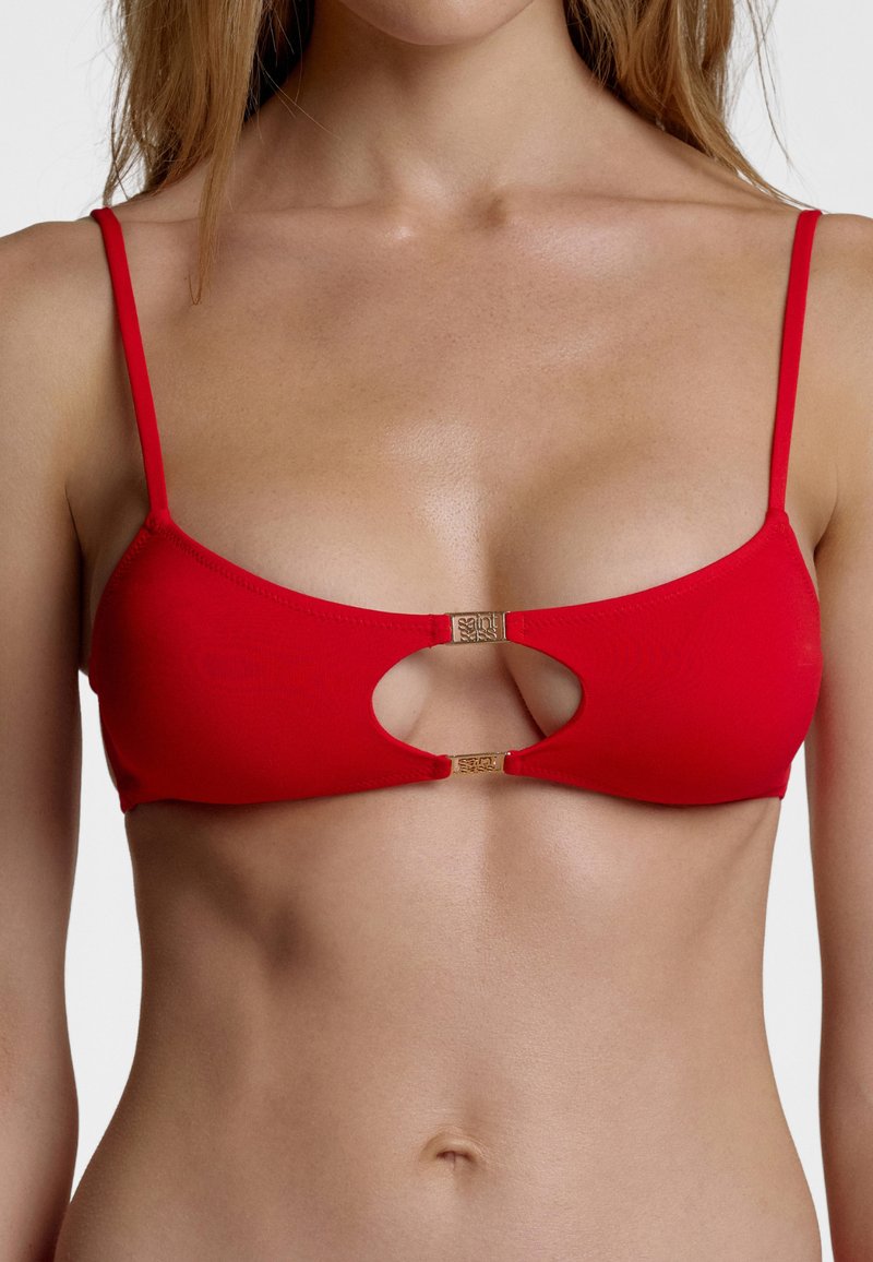 Femme portant un haut de bikini rouge avec des bretelles fines et une découpe centrale maintenue par deux fermoirs dorés.