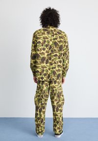 Camo-patterned shirt and pants in green, tan, and brown tones. Loose fit, long sleeves, and cargo pockets on pants. Suitable for casual wear.