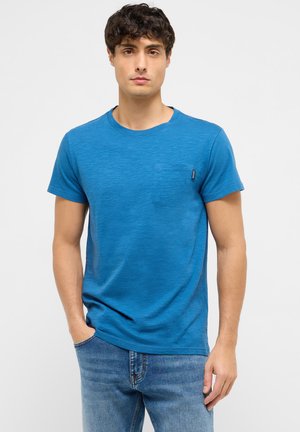 Man with short curly hair wearing a blue crew neck t-shirt with chest pocket and blue jeans, standing against a plain white background.