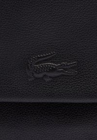 Superficie in pelle nera con una finitura texurizzata, caratterizzata da un logo in rilievo nero a forma di coccodrillo. Design semplice con un dettaglio accentato sottile.