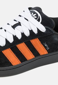 Melns zamšādas sneakers ar baltām auklām, ar trim oranžām svītrām, teksturētām akcentiem un melnu gumijas zoli. Redzams teksts "CAMPUS".