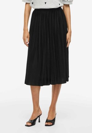 Kobieta ubrana w czarną plisowaną sukienkę midi, szary top z drobnymi czarnymi nadrukami w kształcie serca oraz czarne, odsłaniające palce sandały na obcasie.