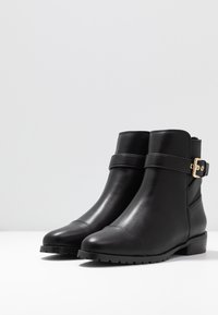Botas de piel negra hasta el tobillo con una textura suave, puntera redonda, herrajes dorados decorativos y una correa ajustable alrededor del tobillo.