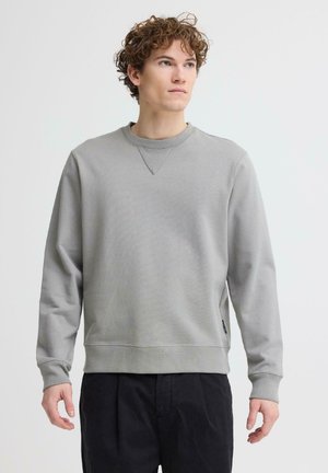 Ung mand med krøllet hår iført en ensfarvet, lys grå crewneck-sweatshirt og sorte bukser, stående mod en hvid baggrund.