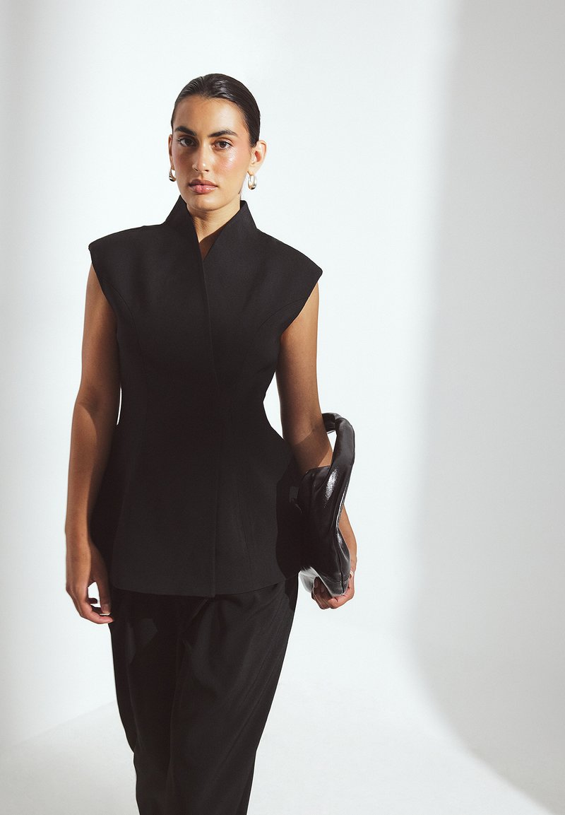 Schwarzes strukturiertes Oberteil mit hohem Kragen und Schulterpolstern, kombiniert mit lockeren schwarzen Hosen und einer glänzenden schwarzen Clutch-Tasche. Glatte Textur.