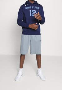 Felpa navy con "GRIZZLIES" e "12" in azzurro chiaro, abbinata a pantaloni corti grigi. Le scarpe da ginnastica sono bianche con il logo swoosh nero.