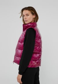 Magenta puffer vest met hoge kraag, gewatteerd patroon, glanzende afwerking en logo aan de onderrand. Gecombineerd met een zwart langemouwen top.