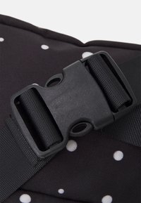 Gros plan d'une boucle en plastique noire à libération latérale fixée à des sangles en nylon noir réglables sur un tissu noir à pois blancs.