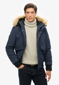 Veste matelassée bleu marine avec capuche en fausse fourrure, poches avant et poignets côtelés, portée sur un pull à col roulé gris clair et un jean foncé.