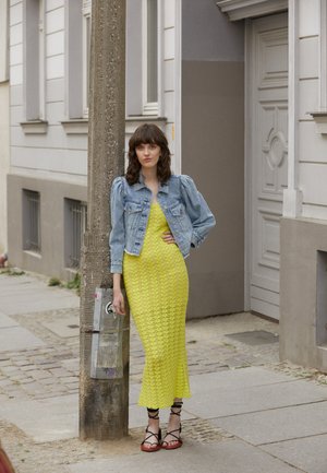 Jaqueta de ganga com mangas bufantes usada por cima de um vestido de malha amarelo brilhante. O conjunto é complementado com sandálias pretas de tiras, com um fundo em coluna.