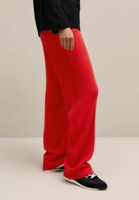 Rode gebreide wijde broek met een zachte textuur, kenmerken een relaxte pasvorm. Gepaard met zwarte sneakers. Achtergrond is neutraal en minimalistisch.
