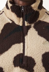 Brun och krämfärgad fleecepullover med halvmanschett, som har ett mönster inspirerat av ko och en texturerad yta. Litet logo på nedre vänstra sidan.