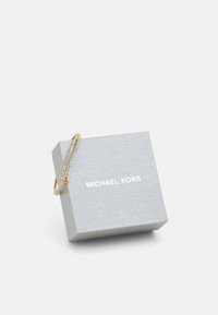 Sidabrinė dovanų dėžutė su tekstūruota paviršiumi, ant kurios yra balta "MICHAEL KORS" logotipas, kartu su auksine grandinėlę apyranke su skaidriais akcentais.