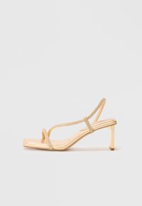 Strappy high-heeled sandal in gold, featuring embellished straps, a square toe design, and a thin block heel for stability.
