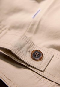 Tessuto beige con una texture liscia, caratterizzato da un bottone marrone scuro con un design a forma di teschio. Dettagli di cucitura lungo i bordi della patta.