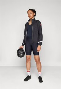 Juoda dviratininko striukė su atspindinčiais akcentais, derinama su juodu bodžiūtu ir šortais. Dėvi juodą šalmą, stovi su dviratininko batais.