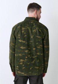 Veste en tissu camouflage dans des nuances de vert et de brun, avec un col à boutons, des coutures sur la poitrine et des manches longues avec des poignets à boutons. La texture semble douce.