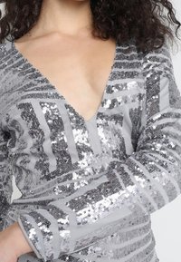Robe en sequins argentés avec décolleté en V profond et manches longues, présentant un motif rayé de sequins réfléchissants sur un tissu gris doux.