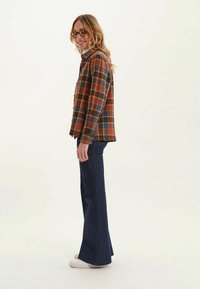 Chemise en flanelle à carreaux aux tons orange, marron et bleu ; associée à un jean évasé bleu foncé et des baskets blanches. Vue de profil.