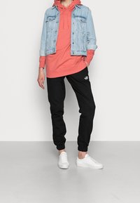 Giacca di jeans di colore blu chiaro, felpa arancione, pantaloni jogger neri con logo e sneaker bianche. Outfit casual con design a strati.