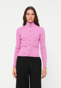 Frau in einem eng anliegenden, rosa gerippten Cardigan mit goldenen Knöpfen und Klappen Taschen, kombiniert mit schwarzen weiten Hosen, vor einem weißen Hintergrund.
