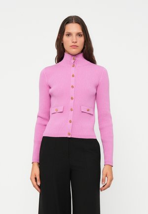 Kvinne iført en tettsittende rosa ribbestrikket cardigan med gullknapper og klaffelommer, kombinert med svarte vide bukser, mot en hvit bakgrunn.