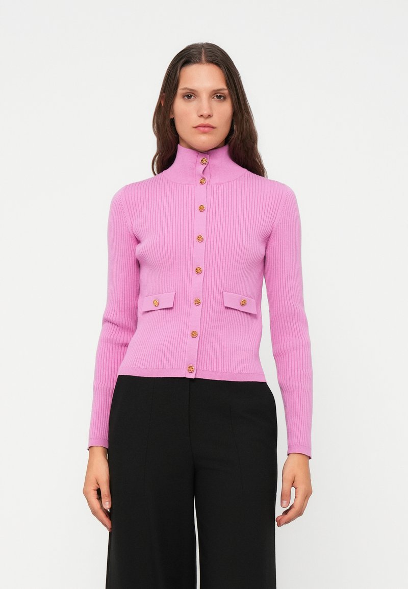 Frau in einem eng anliegenden, rosa gerippten Cardigan mit goldenen Knöpfen und Klappen Taschen, kombiniert mit schwarzen weiten Hosen, vor einem weißen Hintergrund.