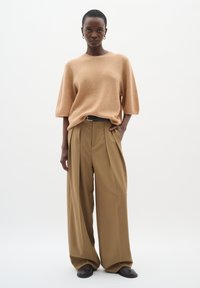 Beige gerippter Kurzarmpullover kombiniert mit hochgeschnittenen, weiten, tanfarbigen Hosen. Das Model trägt einen schwarzen Gürtel und braune Schuhe. Schlichte, klare Linien.