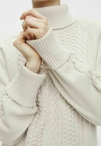 Pull en maille torsadée beige clair, avec un col montant et des poignets côtelés, mettant en valeur des motifs texturés sur le devant et les manches.