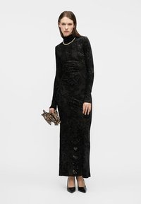 Γυναίκα που φοράει μακρύ μαύρο φόρεμα με σχέδιο, κολιέ από πέρλες, μαύρες γόβες, κρατώντας μια μικρή τσάντα χειρός με λεοπάρ σχέδιο, στέκεται σε λευκό φόντο.