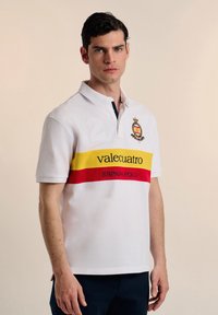 Camisa polo blanca con cuello azul, con un gran número "2", una banda a rayas amarilla y roja, y el texto "valecuatro" y "ESPAÑA POLO."