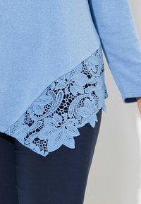 Maglietta azzurro chiaro con una finitura testurizzata, caratterizzata da un orlo in pizzo con motivi floreali. Abbinata a pantaloni blu scuro.