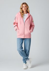 Felpa rosa con zip e tasca frontale, dettagli stampati sulle maniche, abbinata a jeans azzurri e sneakers bianche. Tessuto morbido.
