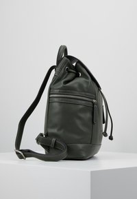 Sac à dos en cuir vert avec une finition texturée, fermeture à cordon, poche zippée sur le côté et bandoulière réglable avec surpiqûres contrastées.