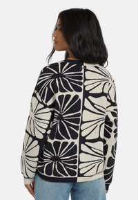 Pull en tricot ivoire avec de grands motifs floraux bleus marine. Il présente un col rond et un ourlet côtelé, avec des épaules tombantes pour un ajustement décontracté.