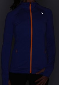 Giacca sportiva blu con cerniera arancione e dettagli in arancione. Presenta maniche lunghe e zip frontale. Tessuto leggero, logo sportivo sul petto.