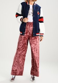 Marineblaues Varsity-Jackett mit weißen Fleeceärmeln und roten Akzenten, weißes Grafik-T-Shirt und pinke Samt-Hosen in weiter Beinform; silberne Schuhe.