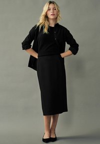 Sort langærmet skjorte med knapper over en sort sweater og en tætsiddende sort midi-nederdel. Tekstureret stof, strømlinet silhuet og ingen synlige mønstre.