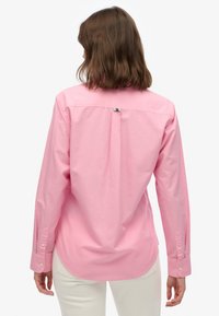 Mujer con cabello castaño hasta los hombros, vistiendo una camisa rosa de manga larga con botones y pantalones blancos, vista desde atrás.