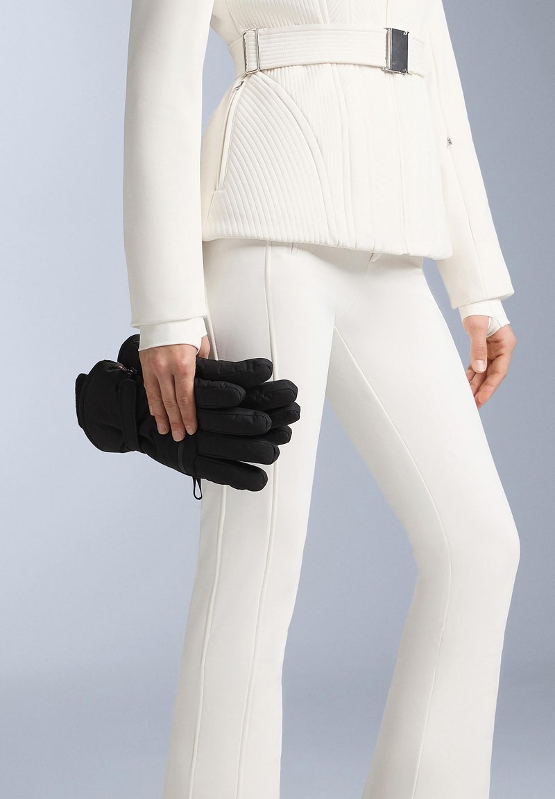 Svarta isolerade handskar med vadderad design och femfingerskonstruktion, hållna avslappnat i en hand, kombinerade med en figursydd vit outfit.