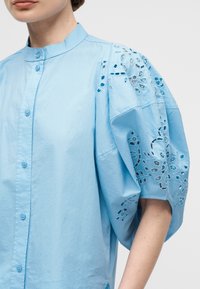 Γυναίκα που φοράει μια γαλάζια μπλούζα με κουμπιά, μανδάριν ζωνάρι και φουσκωτά μανίκια με κεντήματα λουλουδιών σε στυλ eyelet.