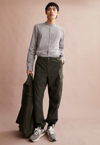 Grijze lange mouwen shirt met een Mandarin kraag, elastische taille donkergroene cargobroek met zakken, en grijze sneakers met witte accenten.