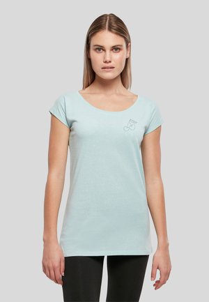 Jeune femme portant un t-shirt bleu clair à manches courtes avec un petit motif de cerise sur la poitrine gauche, debout devant un fond gris.