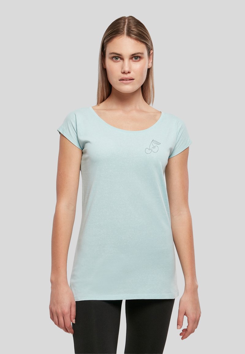 Jeune femme portant un t-shirt bleu clair à manches courtes avec un petit motif de cerise sur la poitrine gauche, debout devant un fond gris.