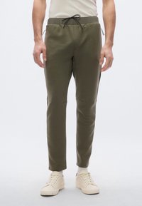 Oliivinvihreät juoksuhousut, joissa on joustava vyötärönauha ja nyöri. Housuissa on kapeat lahkeet ja minimalistinen muotoilu, ja ne on yhdistetty vaaleisiin lenkkareihin.